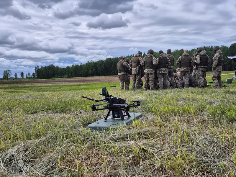 Šauliai susibūrę žaliame lauke nugaromis į kamerą, priekyje sunkusis multirotorius ant tūpimo aikštelės