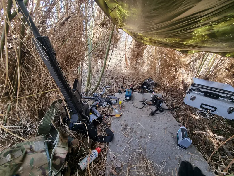 Maskuota slėptuvė po tentu ir nendrėmis: šautuvas, keli FPV dronai ir įranga ant kilimėlio