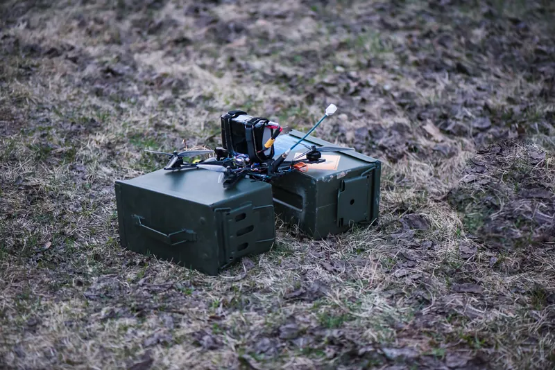 FPV dronas su antena ant dviejų žalių šovinių dėžių purvinoje žolėje