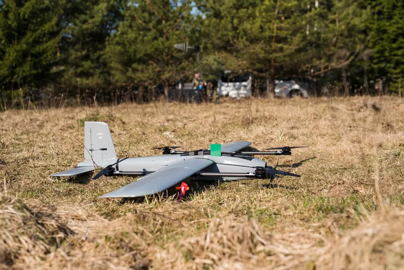 Pilkas VTOL fiksuoto sparno dronas ant sausos žolės, už jo pušys ir automobiliai