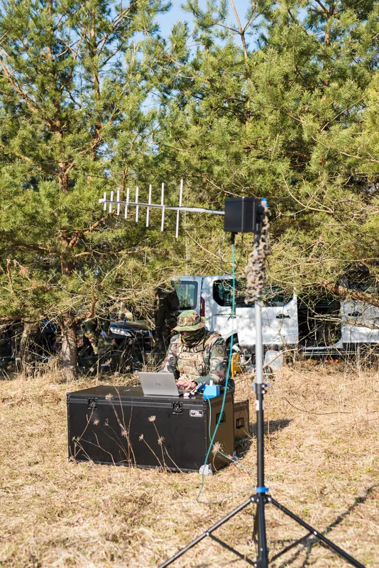 Yagi antena ant trikojo stiebo; operatorius dirba kompiuteriu ant transportavimo dėžės tarp pušų