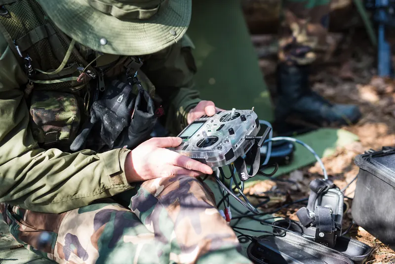 Operatorius su skrybėle, veidas paslėptas po kraštu, ruošia FPV valdymo pultą tarp įrangos