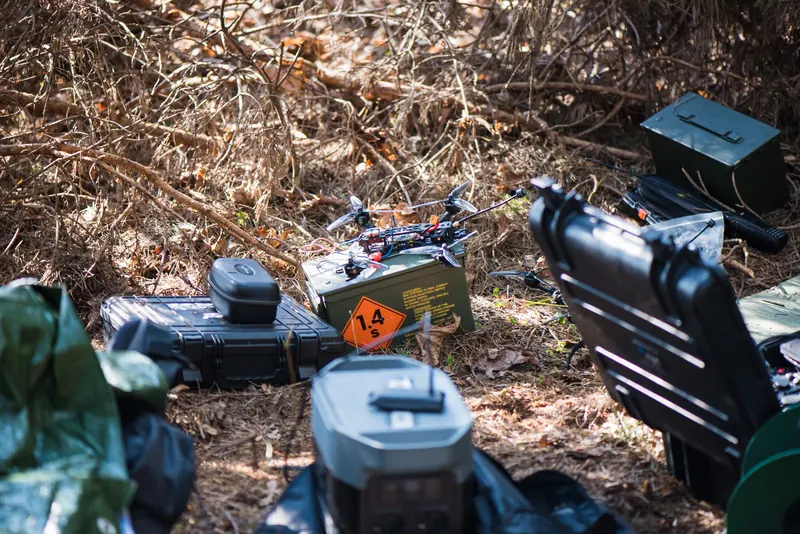 FPV dronas ant šovinių dėžės, šalia kietos dėžės ir akumuliatorius miško paklotėje
