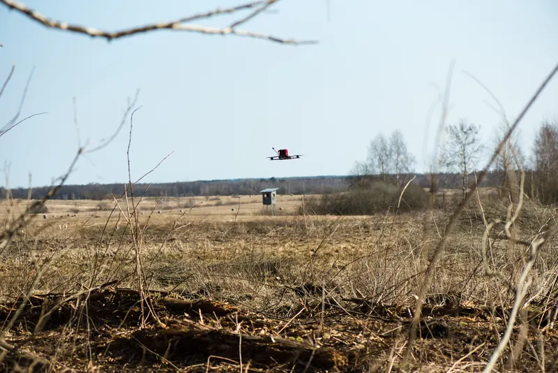 FPV dronas su kroviniu skrenda virš sauso lauko, fone medinis stebėjimo bokštas