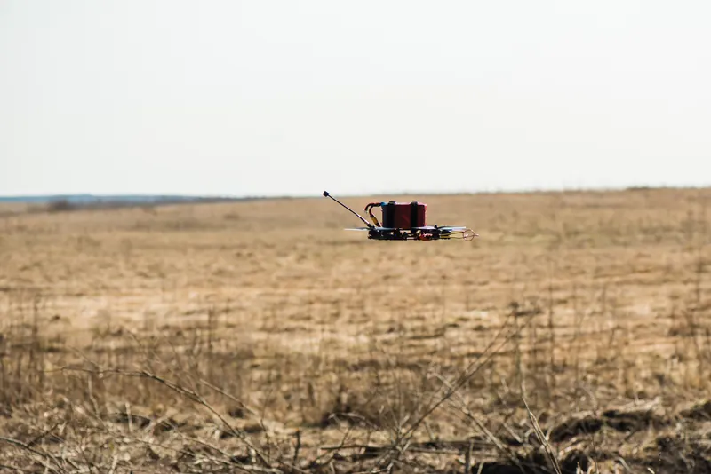 FPV dronas su kroviniu kybo žemai virš lygaus sauso lauko