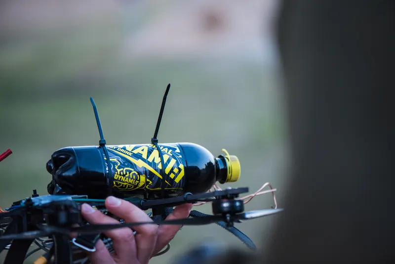 Ranka laiko FPV droną su pritvirtintu energinio gėrimo buteliu kaip bandomuoju kroviniu