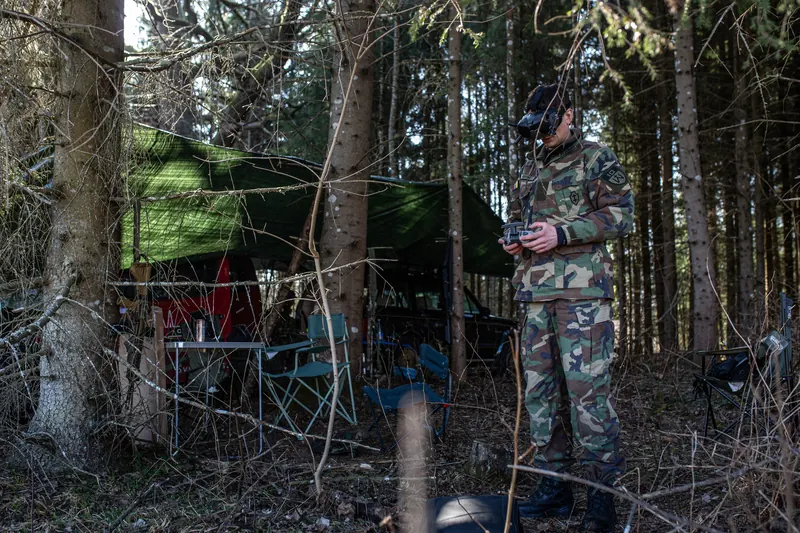 FPV pilotas su akiniais eglyne, rankose pultas, fone stovyklos tentas ir kėdės