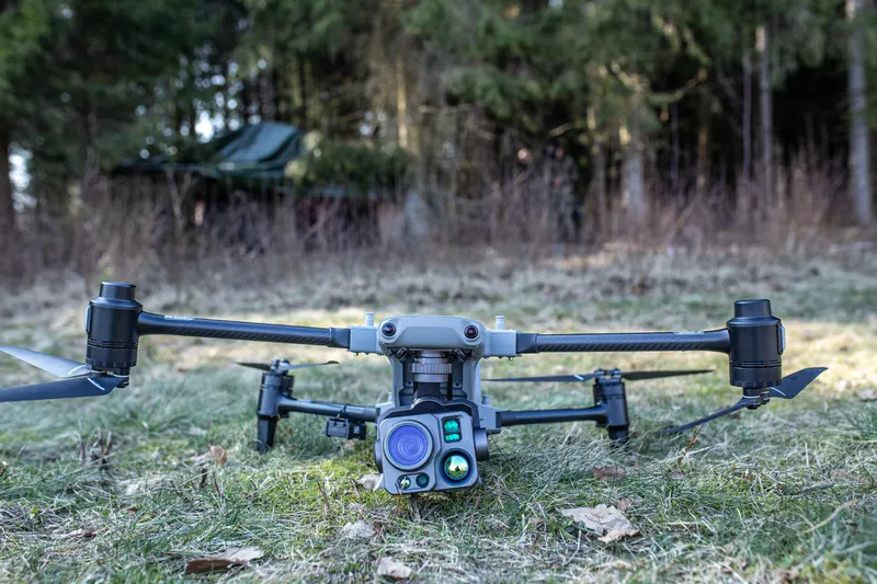 DJI Matrice klasės kvadrokopteris su kamera ant apšalusios žolės, fone miškas