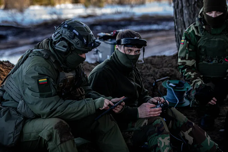 Trys operatoriai sniegingo miško slėptuvėje su FPV pultais ir šviesolaidžio rite