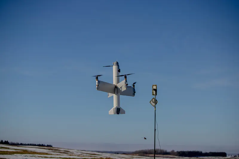 Vertikalaus kilimo dronas kybo giliai mėlyname danguje šalia antenos stiebo, apačioje sniegingas laukas