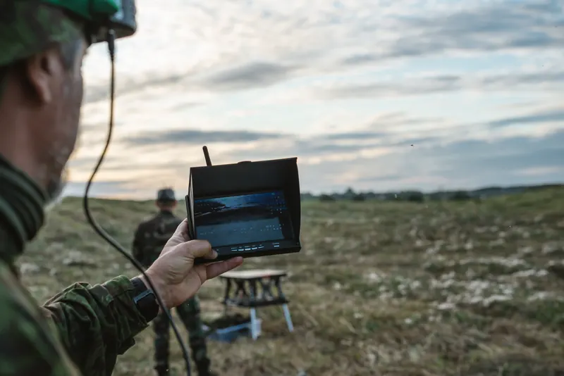 Operatorius laiko FPV monitorių iškėlęs link lauko, fone kitas šaulys ir stalas