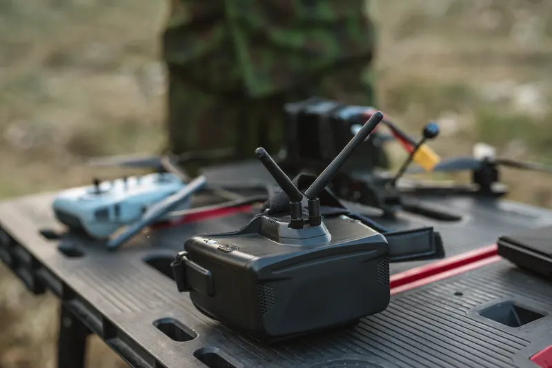 FPV akiniai su antenomis ir pultas ant juodo darbastalio, fone neryški figūra