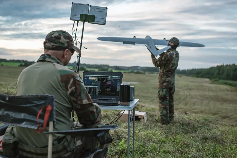 Operatorius prie kompiuterio po paneline antena, kol šaulys iš rankos paleidžia fiksuoto sparno droną