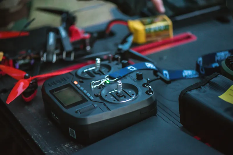 FPV valdymo pultas, kvadrokopteris su raudonais propeleriais ir baterija ant juodo darbastalio
