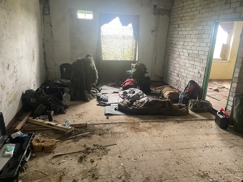 Apleistas kambarys su maskuojamuoju tinklu ant lango, kampe sėdi šaulys, ant grindų įranga ir kilimėliai