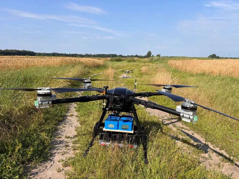 Sunkusis multikopteris su mėlynomis krovinio dėžėmis ant vieškelio tarp kviečių laukų, už jo daugiau dronų